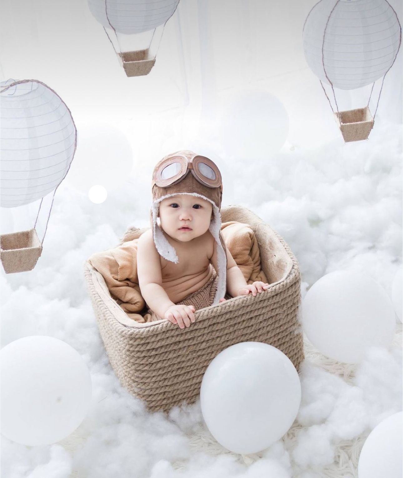 A baby in a knitted aviator hat sits in a woven basket surrounded by white paper lantern hot air balloon decorations.