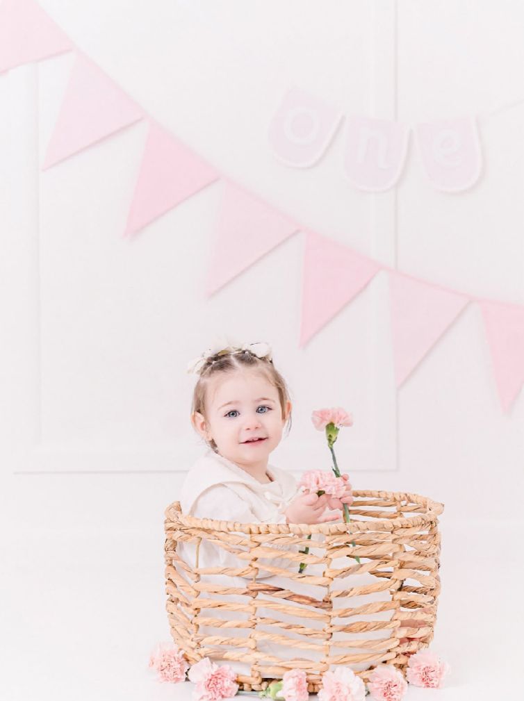 Sweet baby sits in woven basket with pink carnations and bunting banner decorations for spring photo session.