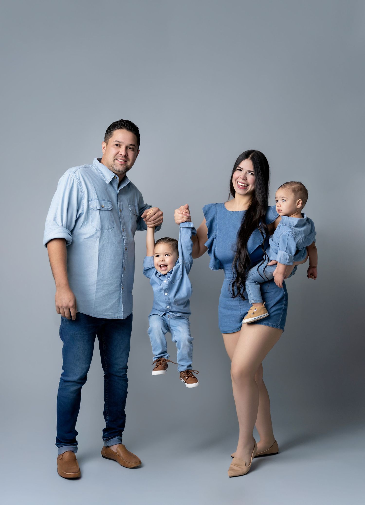 Family of four in matching denim outfits posing in a studio, parents swinging toddler while holding baby.