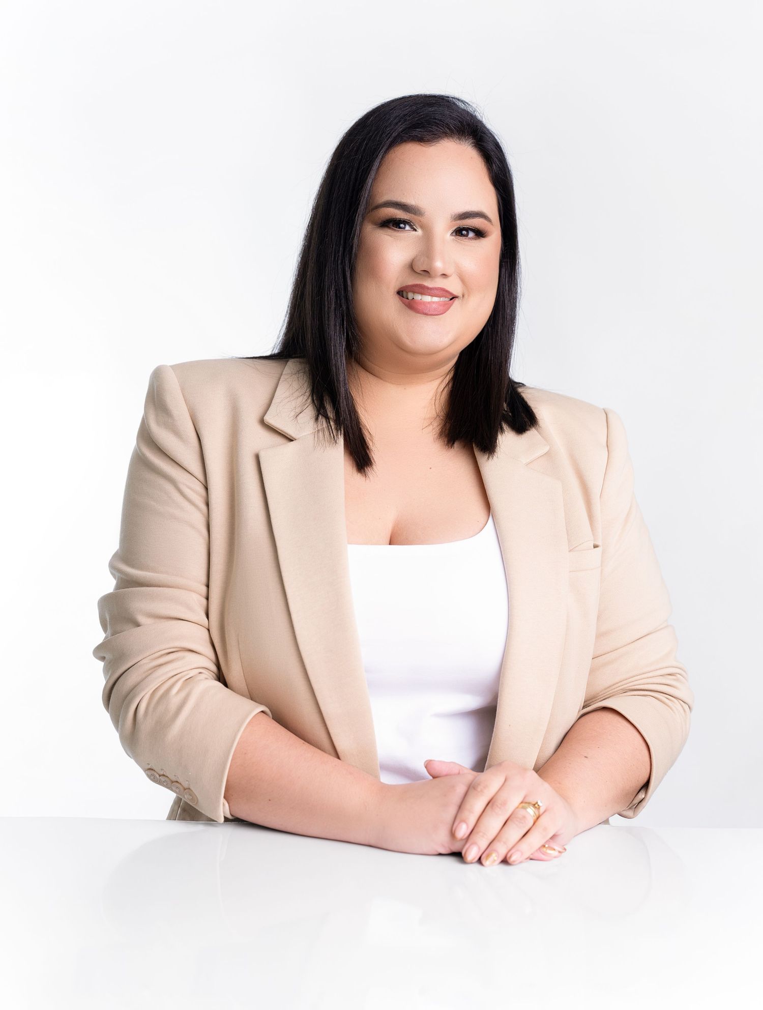 Smiling woman with long dark hair in a beige blazer and white top sits at a white desk with her hands clasped in a professional portrait.