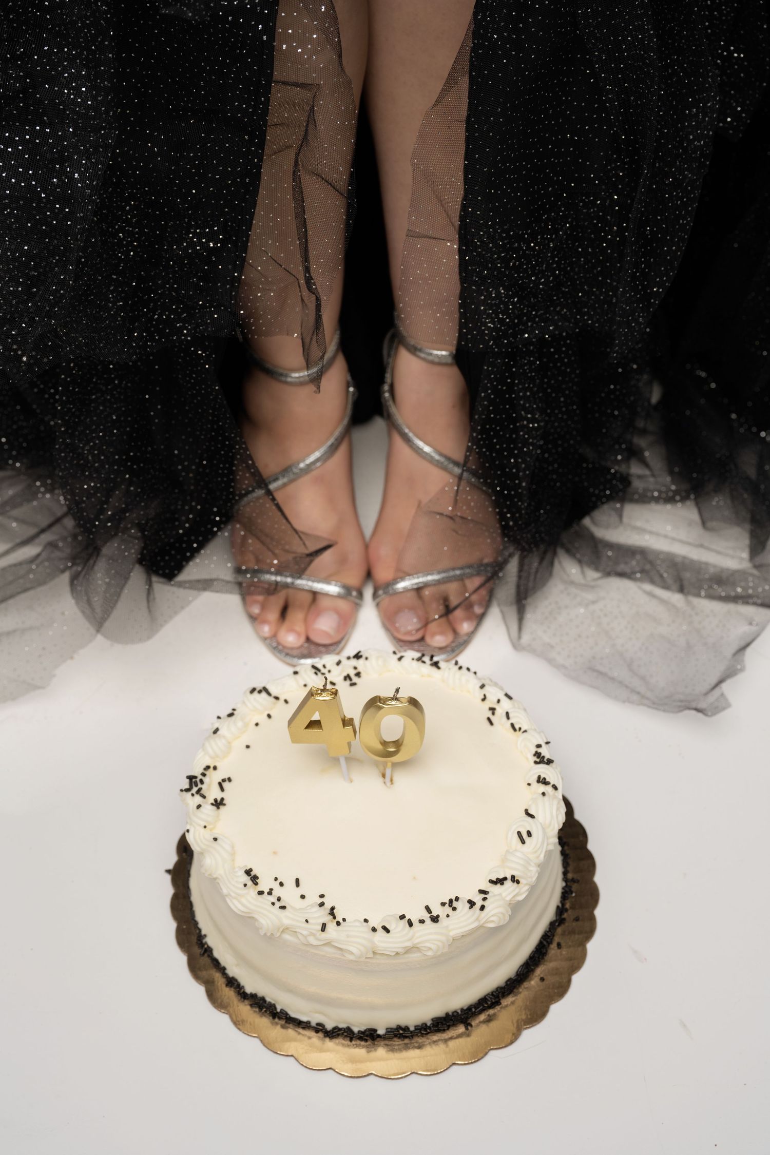 Person in black tulle skirt and silver heels standing beside a white 40th birthday cake with gold candles.