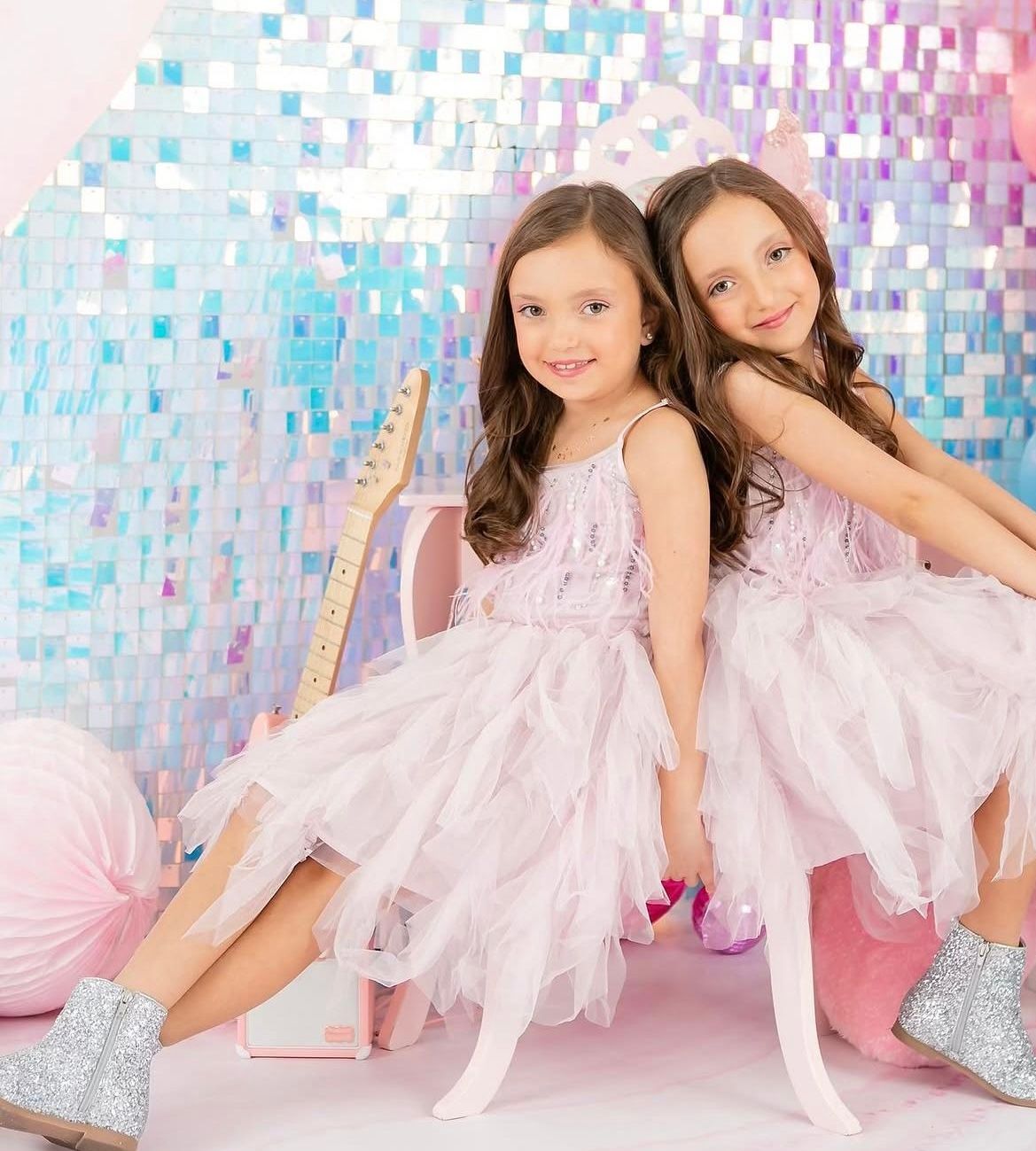 Two young girls in pink tulle dresses and silver boots pose together against a shimmery iridescent backdrop.