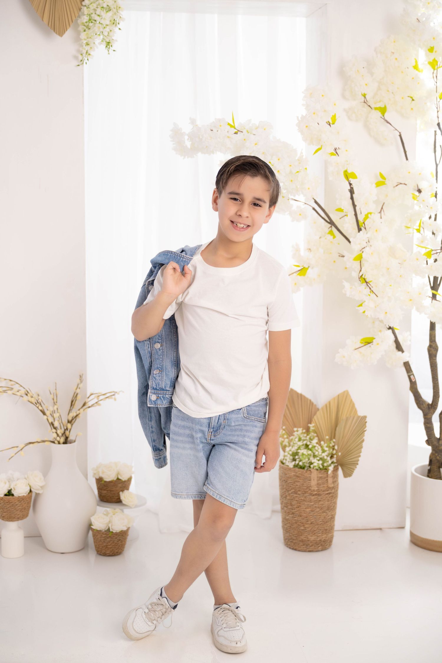 Smiling boy in white tee, denim shorts, holding jacket over shoulder, posing in bright white room with flowers.