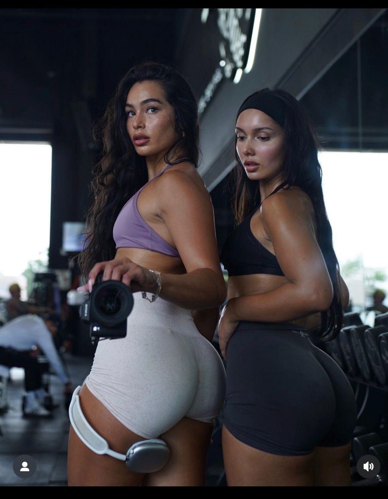 Two athletic women in workout attire pose back-to-back in a gym holding dumbbells.