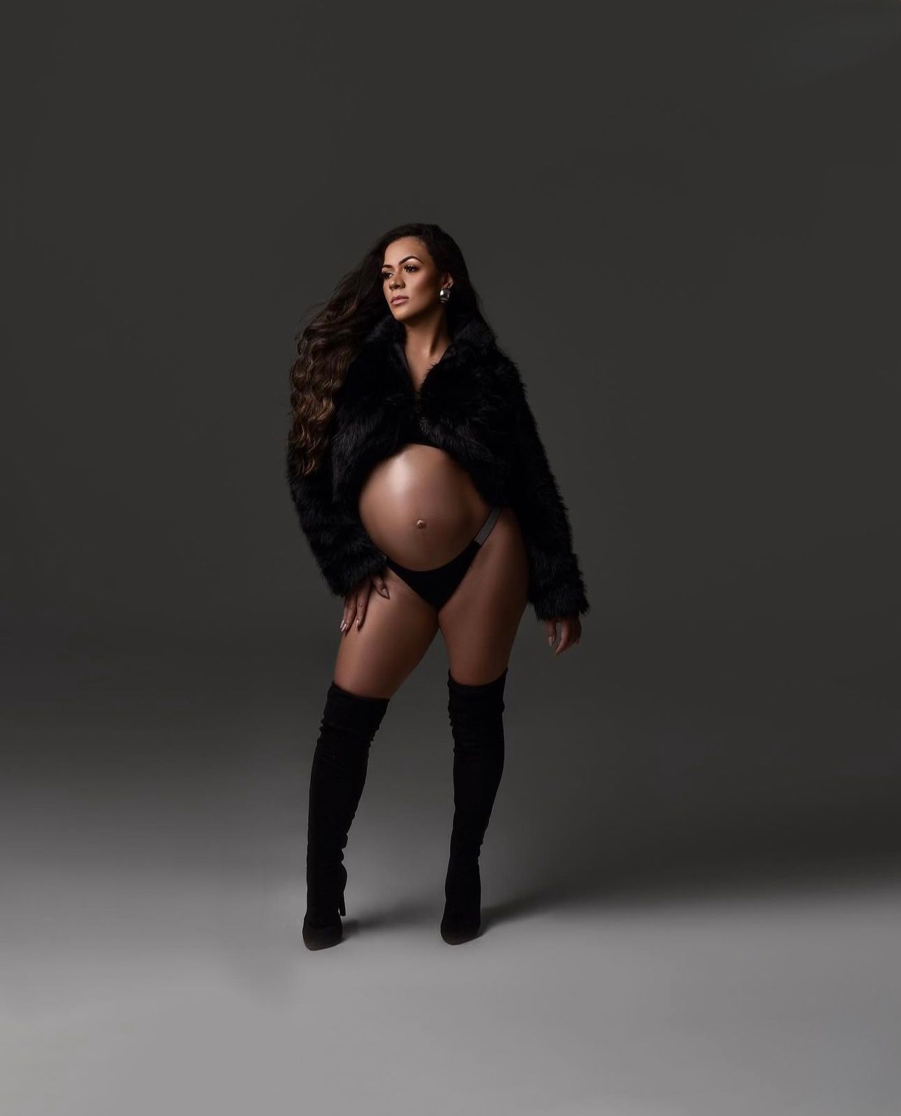 Pregnant woman in black fur coat, thigh-high boots, and bikini bottoms posing confidently in a studio maternity shoot.
