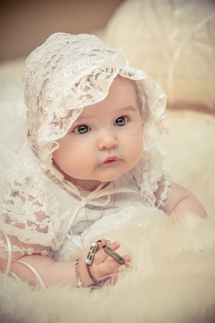 Adorable baby in white lace bonnet and dress lying on fluffy white fur, holding a bracelet, looking at camera.