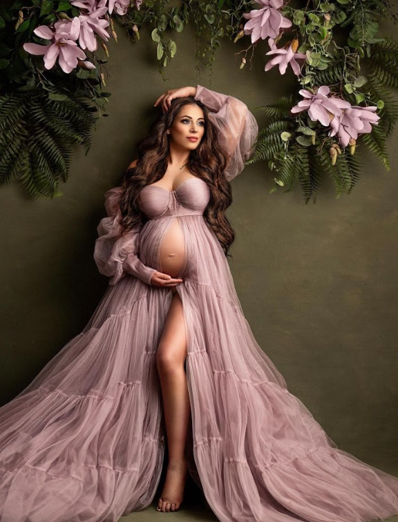 Elegant maternity portrait featuring a flowing mauve tulle gown against a floral backdrop with pink blooms and greenery.