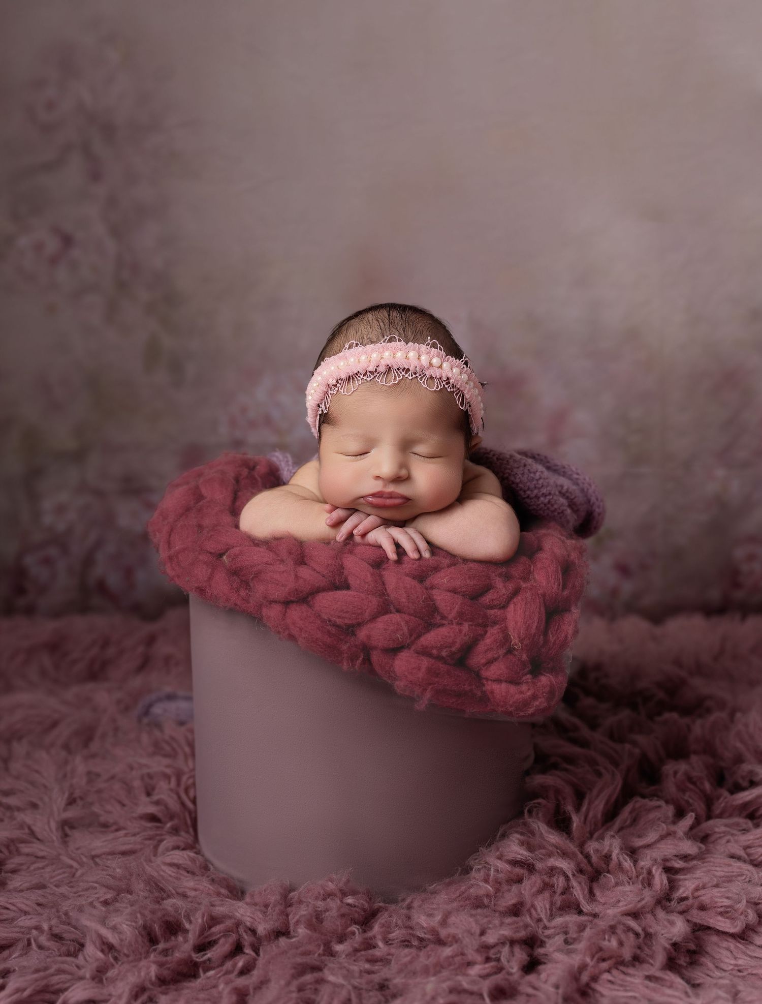 Sleeping newborn baby girl in pink headband posed in mauve bucket with chunky knit blanket on floral backdrop.