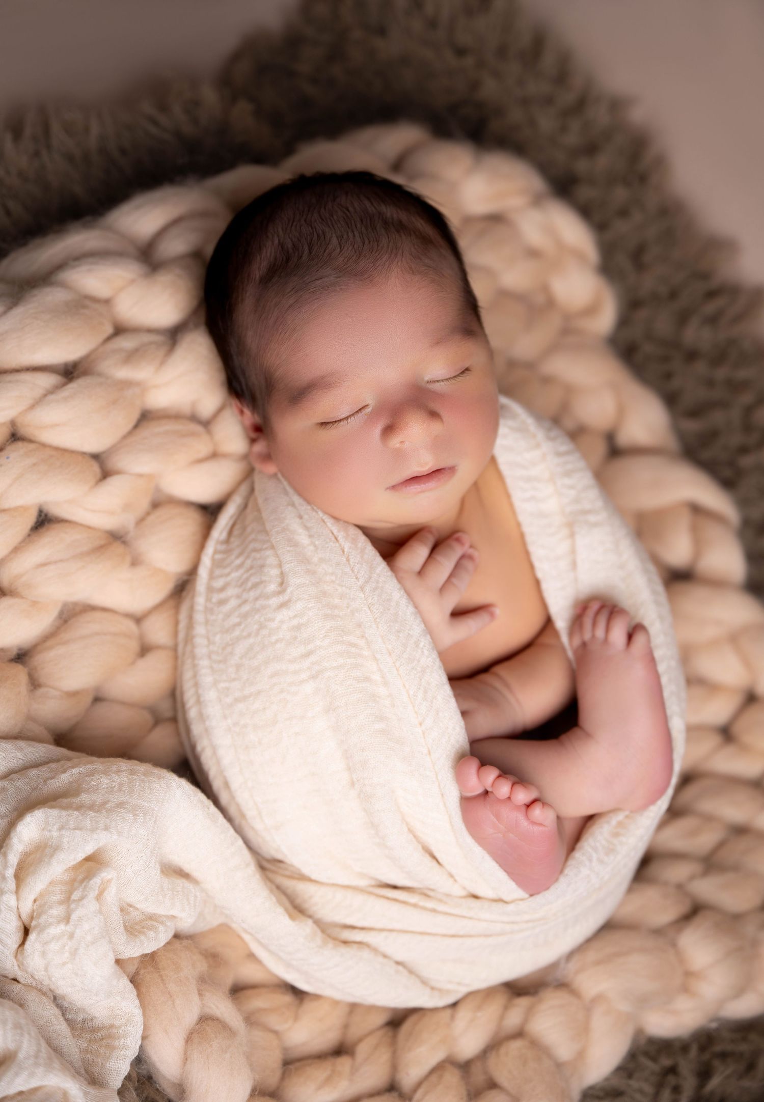 Peaceful newborn baby sleeping wrapped in cream knit blanket nestled in chunky wool basket for professional newborn photography.