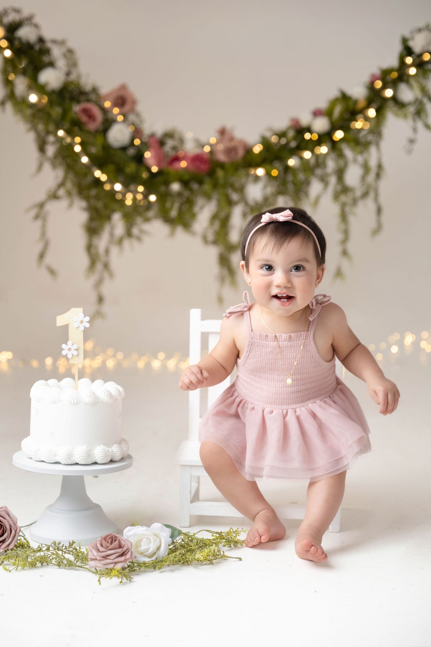 Baby girl in pink dress smiles beside first birthday cake with floral garland and fairy lights backdrop.
