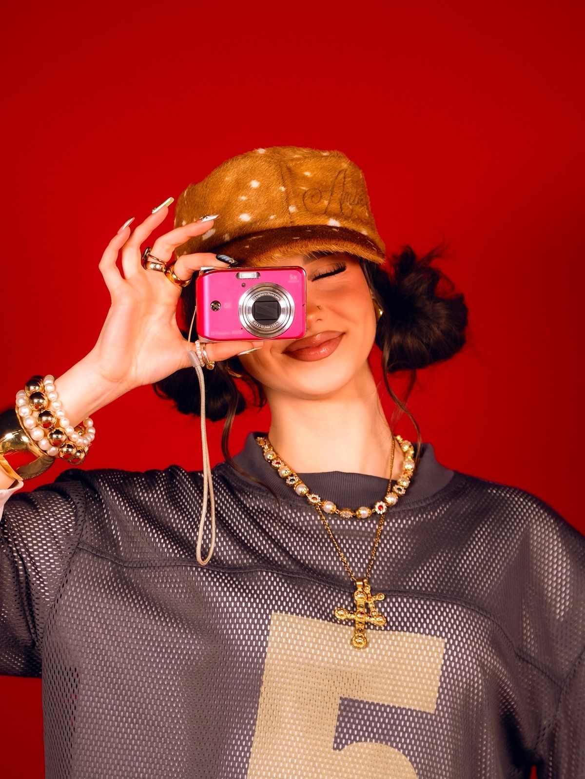 Young woman in brown hat holds pink digital camera, wearing mesh jersey and gold accessories on red background.