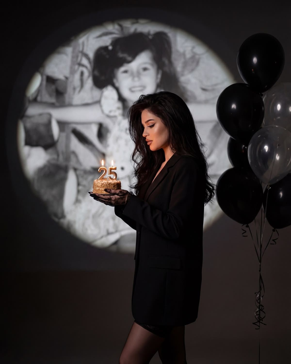 Woman in black dress holds a 25th birthday cake with candles, black balloons and vintage childhood photo projection behind her.