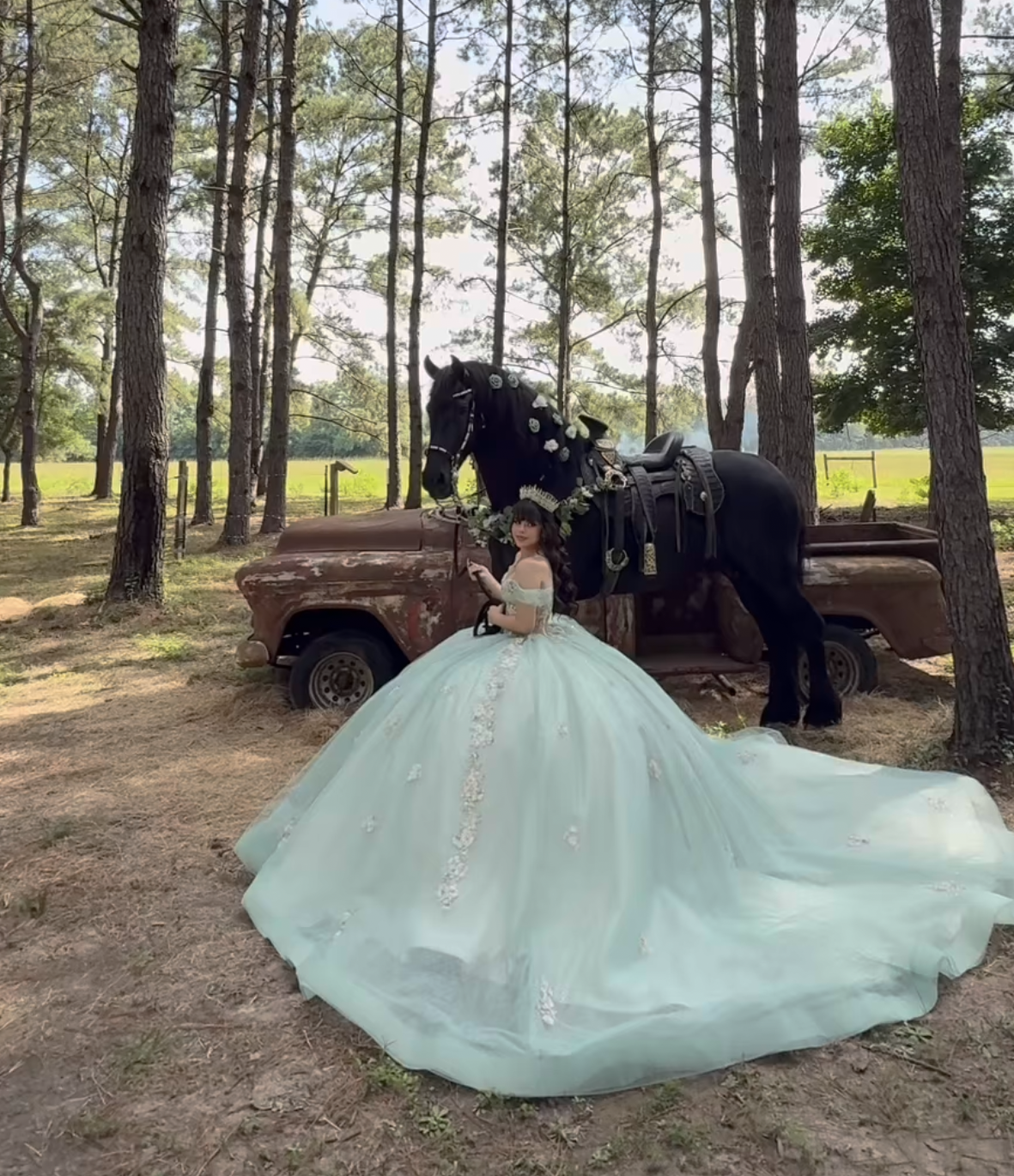 A young woman in a mint green ball gown poses with a black horse near a rustic truck among pine trees.