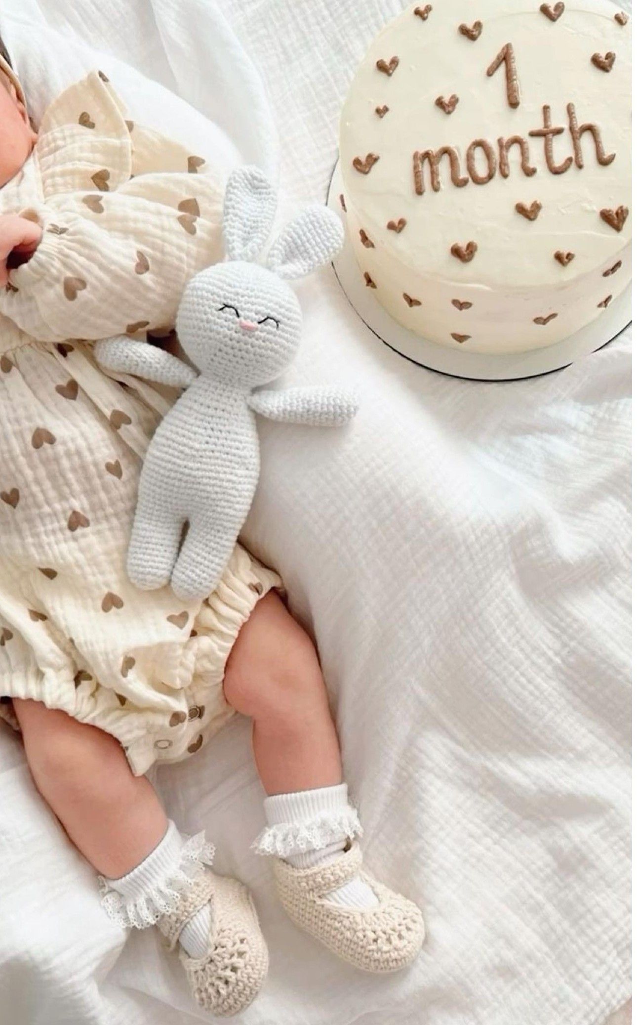 One-month-old baby in heart-print outfit with knit bunny toy and decorative milestone ball on white bedding.