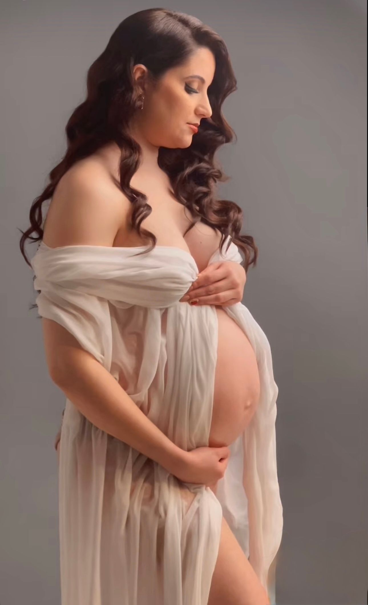 Pregnant woman in sheer white wrap gown cradling her baby bump during an elegant maternity photo shoot.