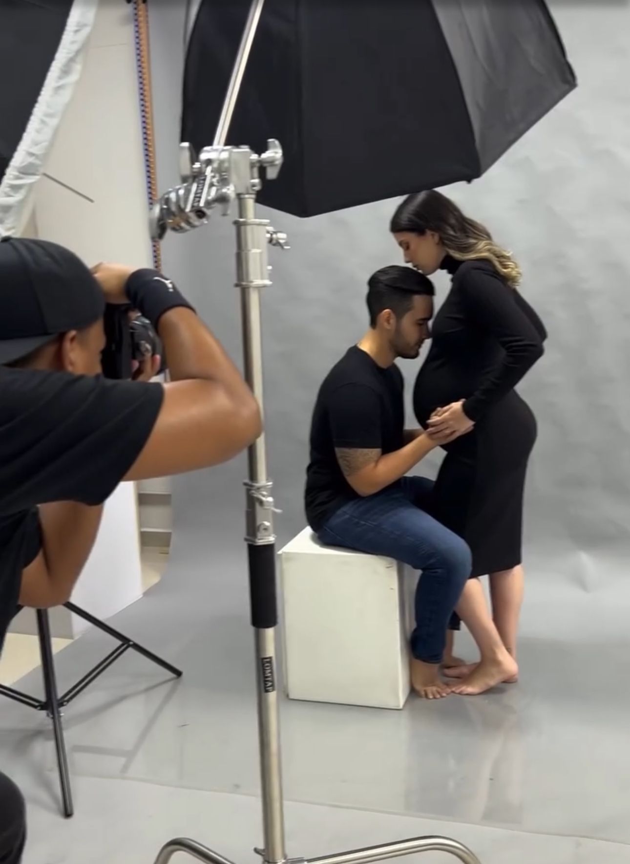 Maternity photoshoot in studio with couple posing while photographer captures the moment under umbrella lighting.