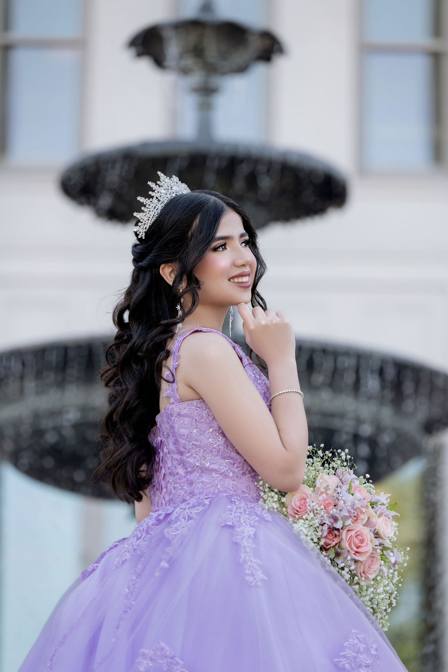 Young Latina in lavender quinceañera ball gown with tiara holds pink bouquet by ornate fountain.