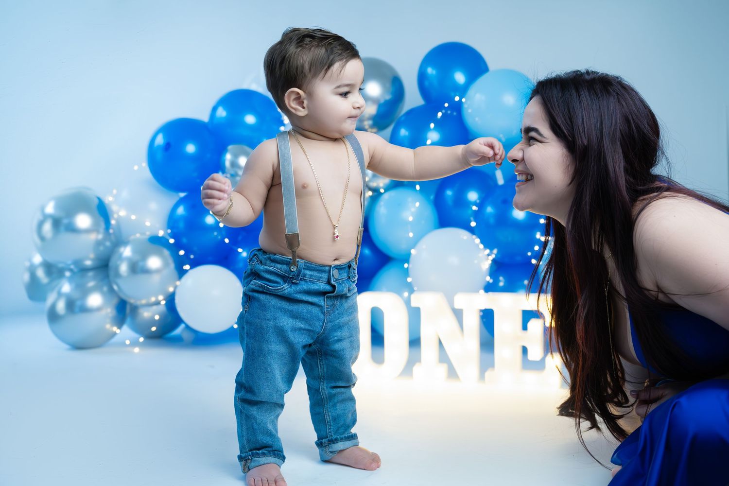 A baby boy in suspenders stands near a ONE sign with blue balloons as his mother smiles at him.