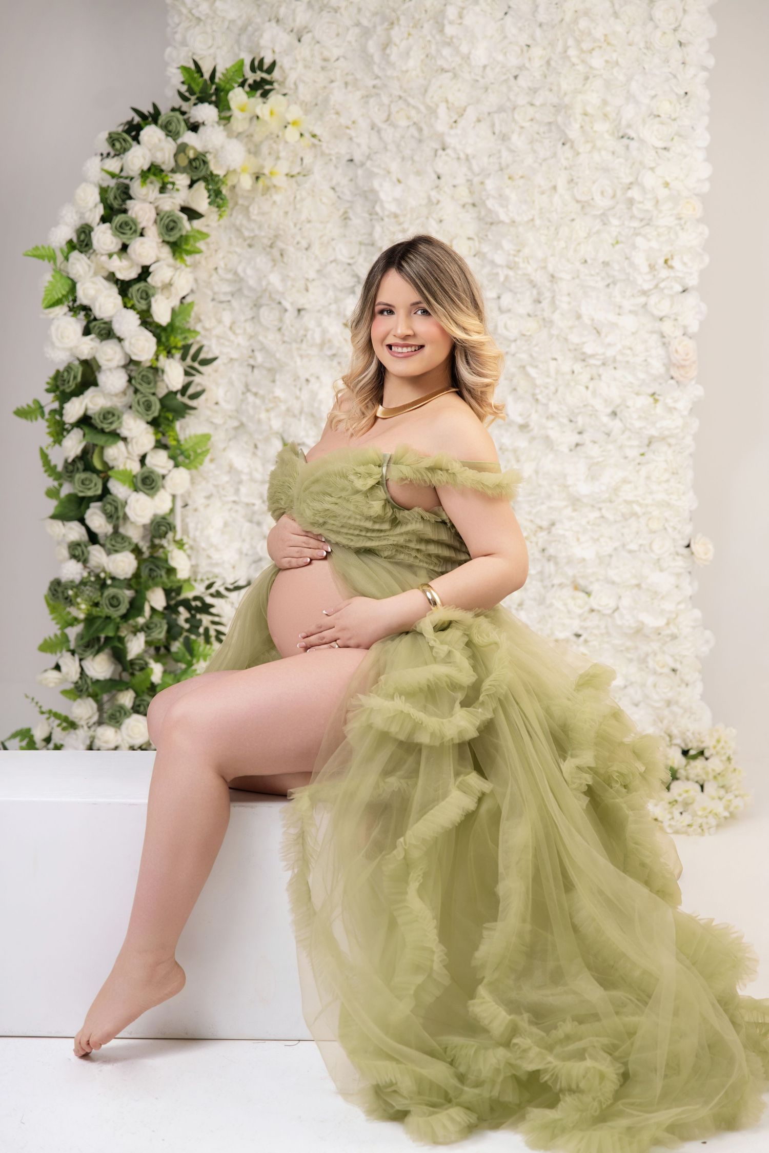 Pregnant woman in olive green tulle maternity gown sitting before white floral wall backdrop.