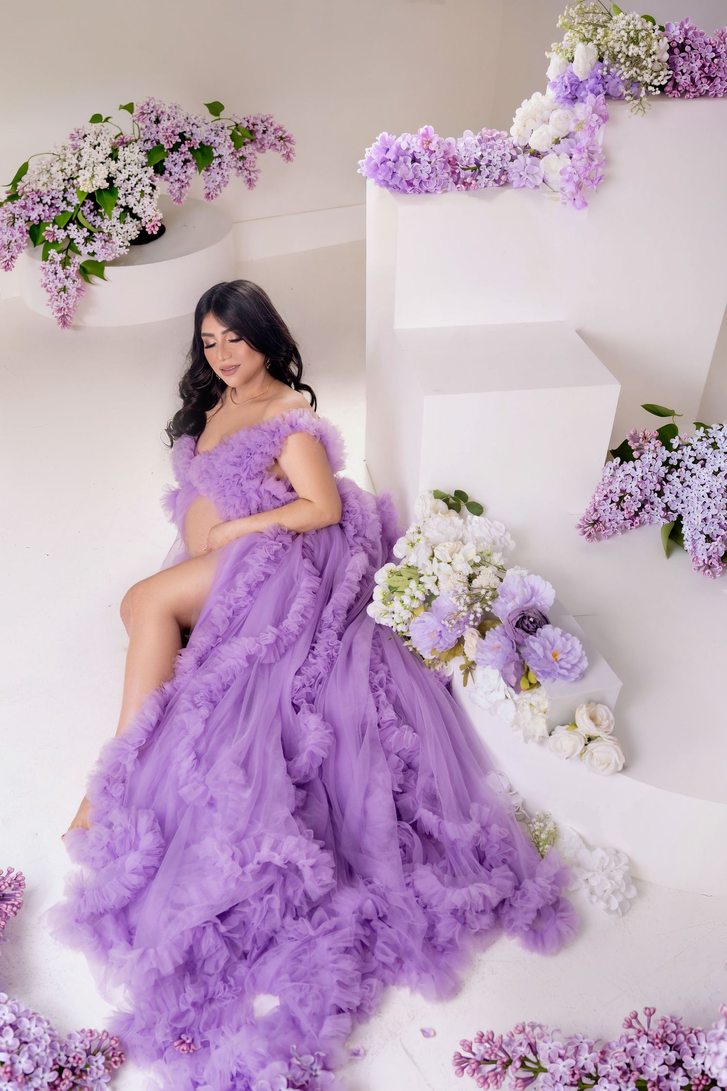 Elegant woman in a flowing purple tulle gown surrounded by lilac flowers in a white studio setting.