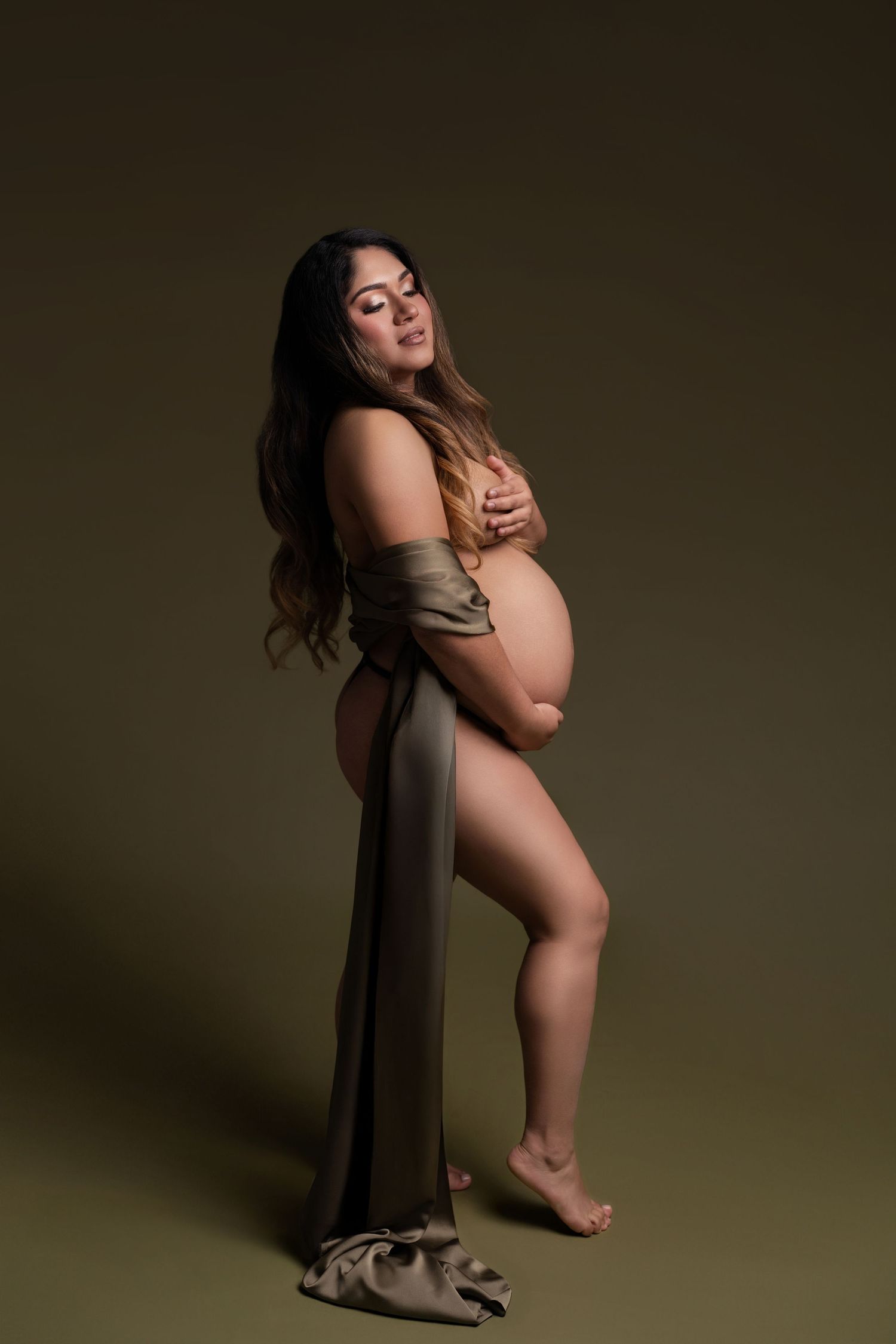 Pregnant woman in olive green draped fabric poses gracefully in studio maternity photography session.