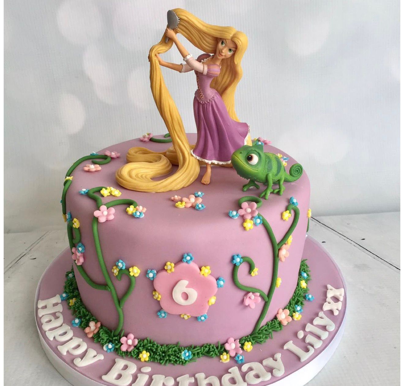 Rapunzel themed birthday cake with Pascal the chameleon, floral decorations, and Happy Birthday Lily writing on pink fondant.