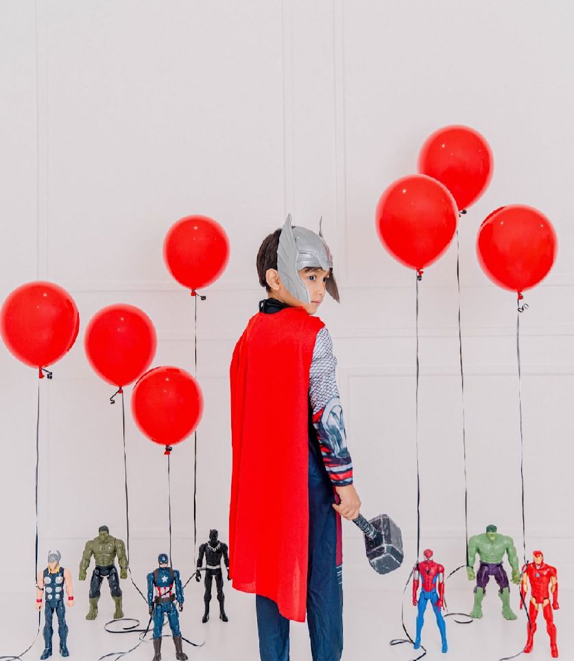 A figure in a red cape stands among superhero action figures with red balloons against a white background.