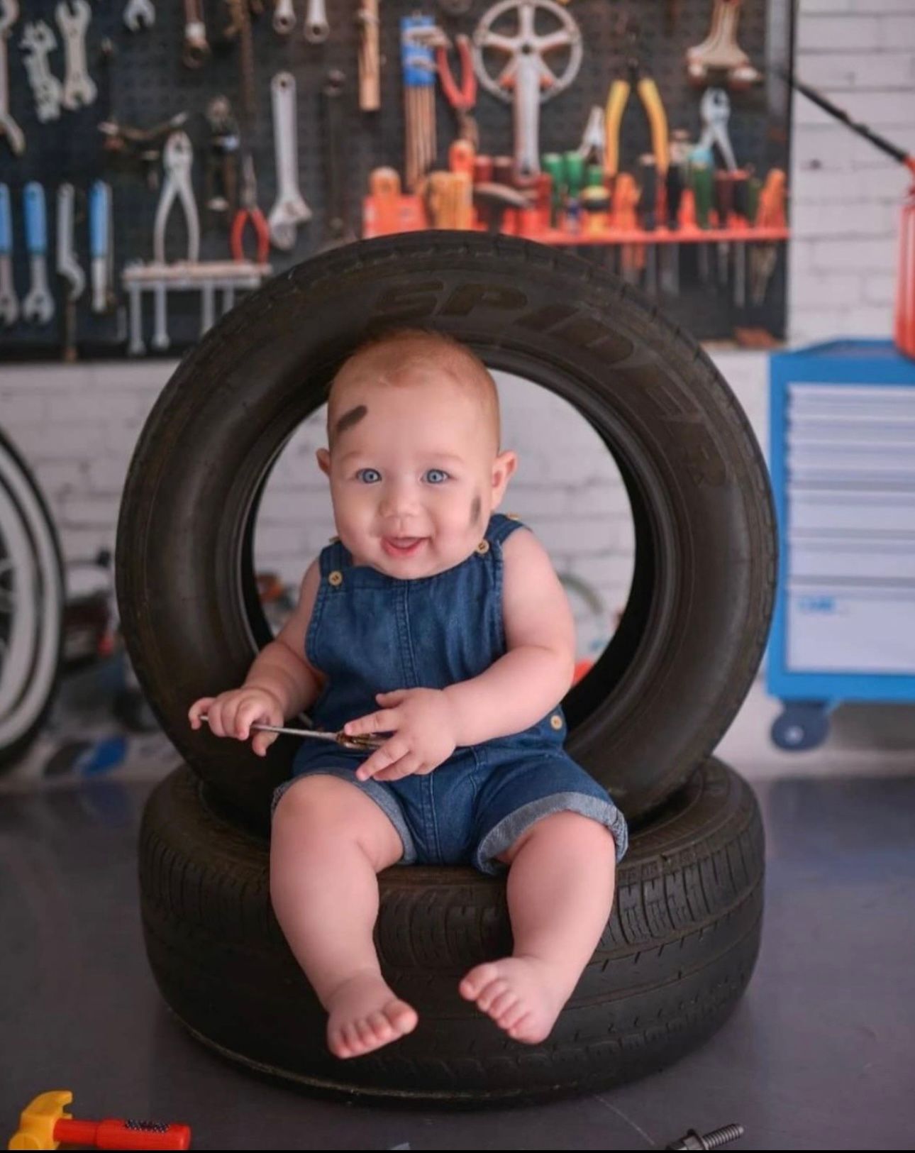Baby in denim overalls sits and plays in a large tire in what appears to be an auto repair shop setting.