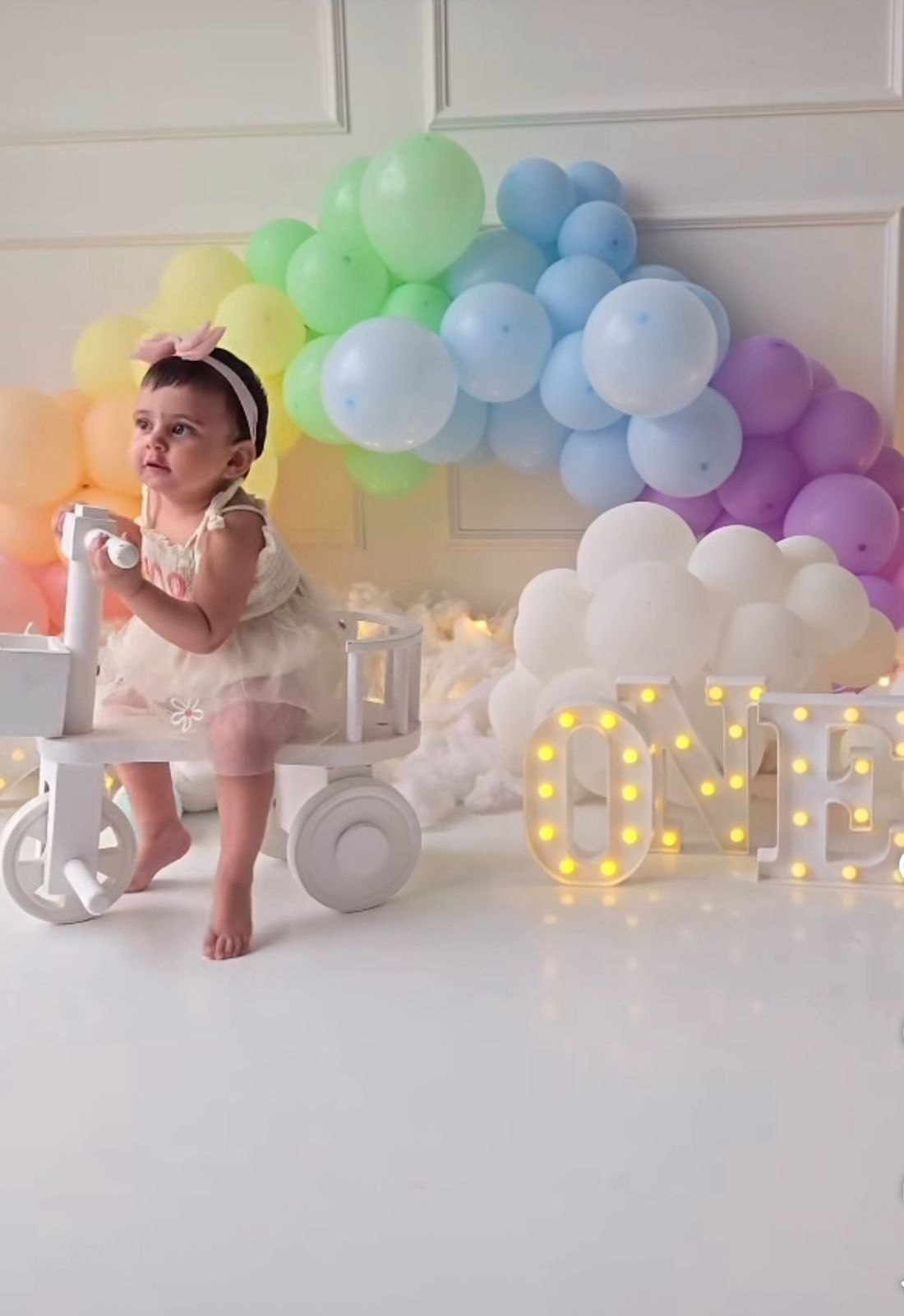 A baby sits in front of a pastel rainbow balloon arch with illuminated letters creating a dreamy photo setup.