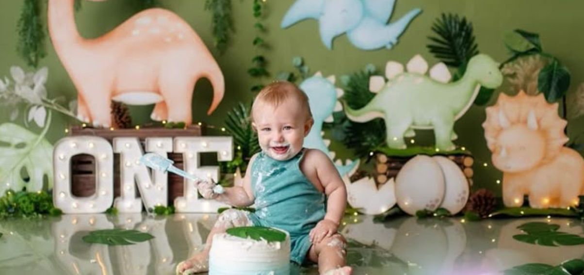 Dinosaur themed celebration backdrop with green foliage and pastel balloon decorations.