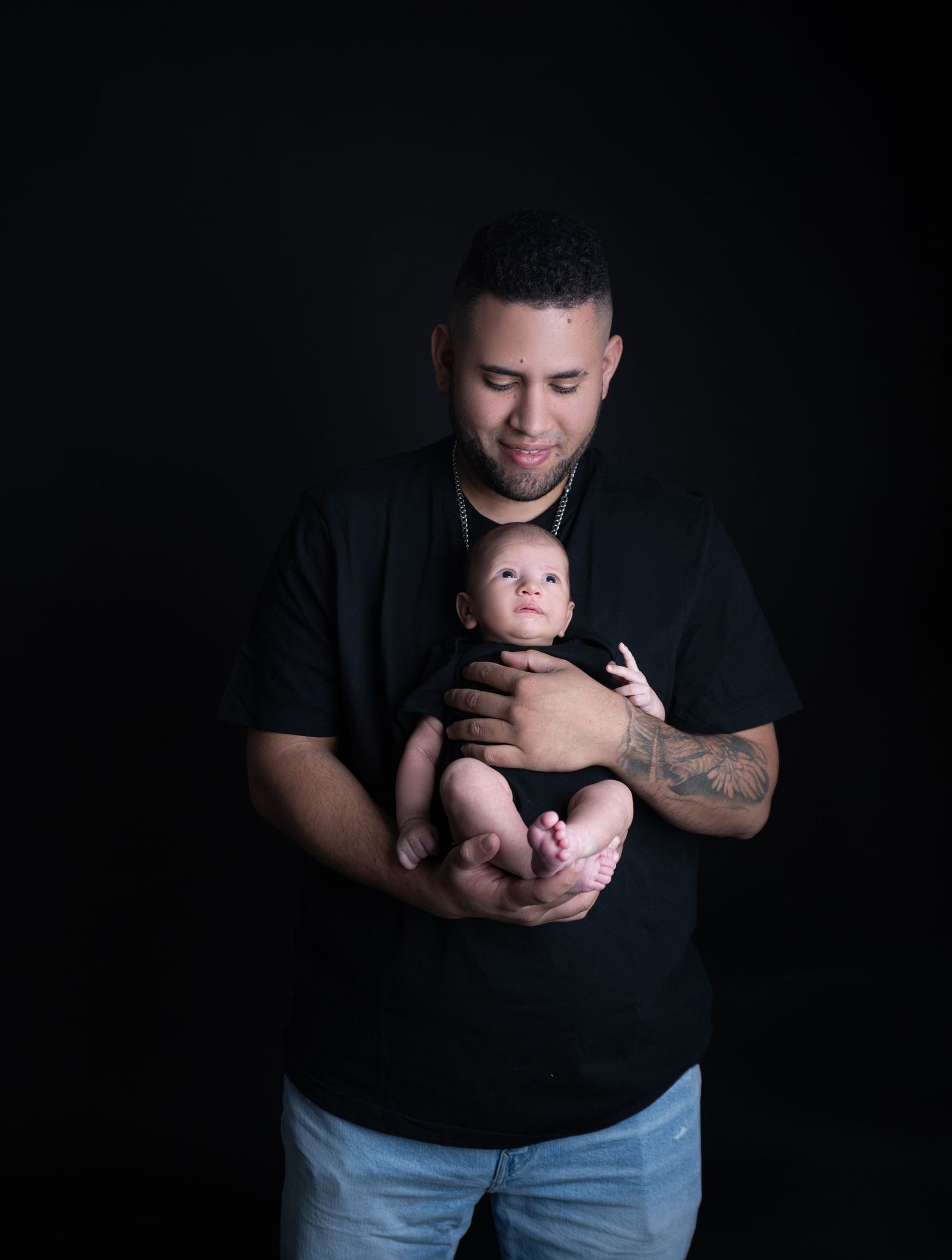 A man in a black t-shirt tenderly holds a newborn baby against a dark studio background.