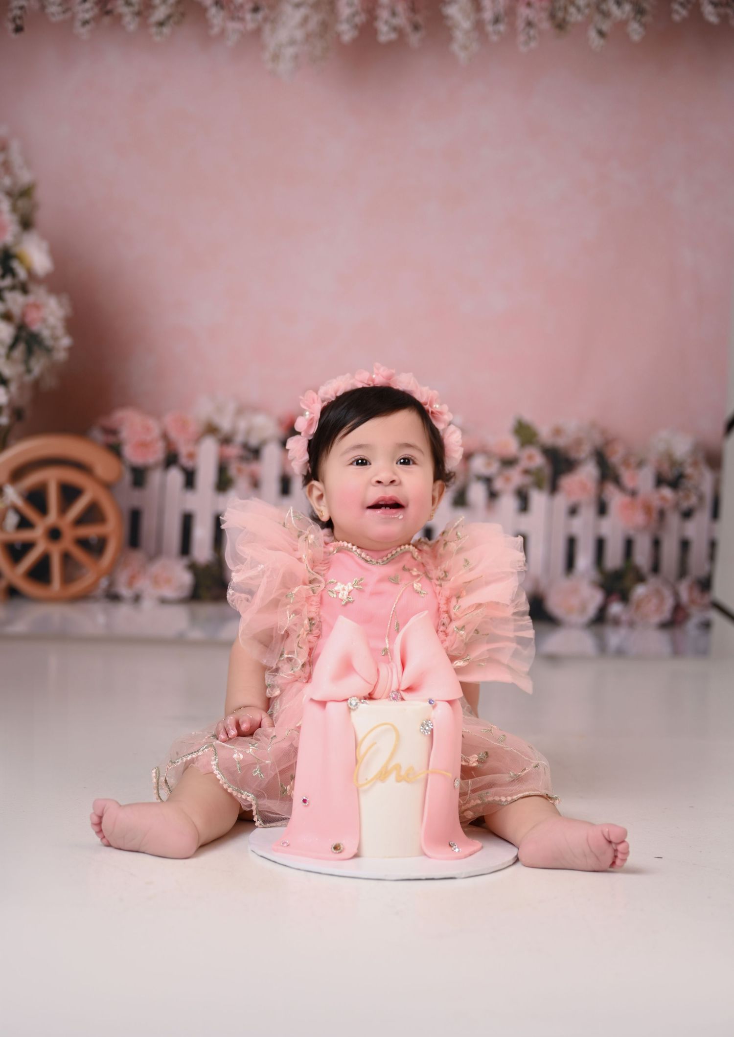 Baby girl in pink dress sits beside a pink ‘One’ cake during a first birthday cake smash photo session.