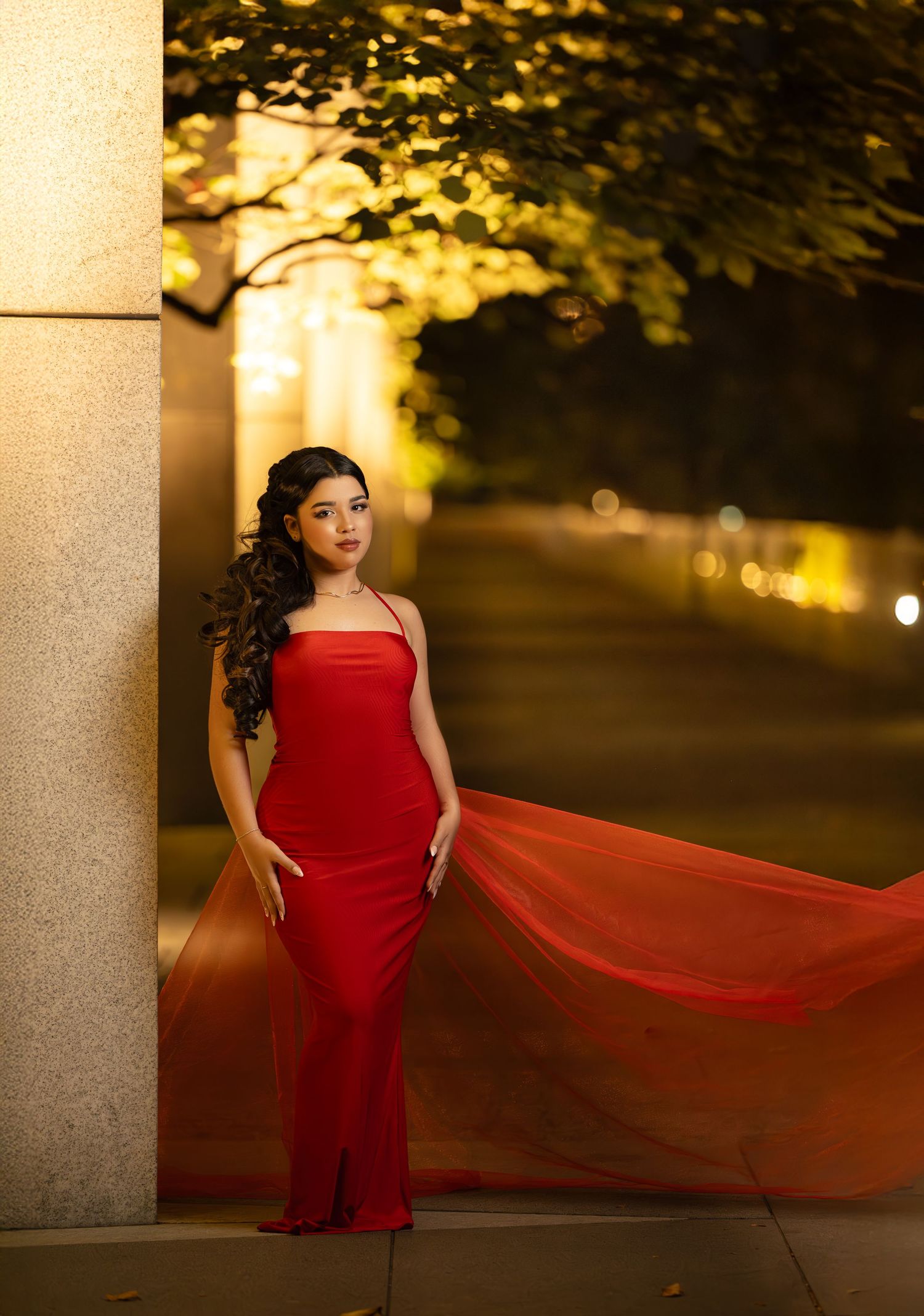 Young woman in elegant red gown poses gracefully against a stone column at night with bokeh lights behind her.