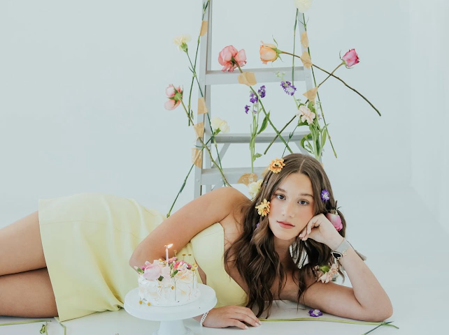 Young woman in yellow dress lying beside floral birthday cake with spring flowers.