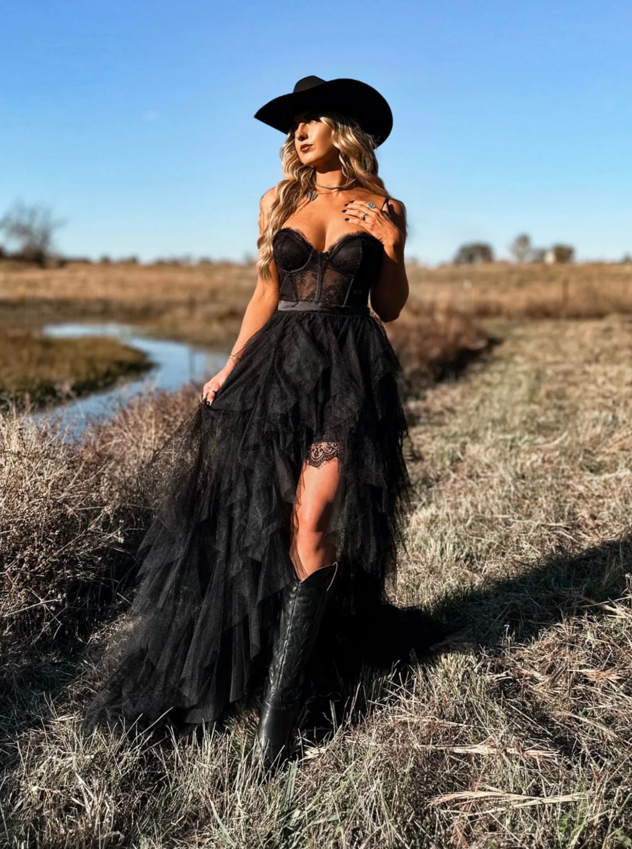 Woman in black lace corset gown, cowboy hat, and boots posing in a rural field at golden hour.