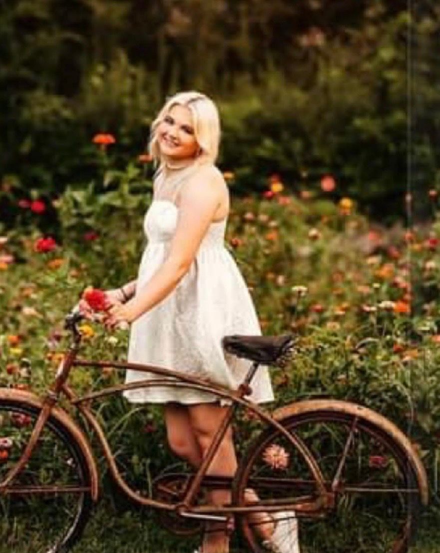 Young blonde woman in white dress posing with vintage rusty bicycle in colorful flower garden.