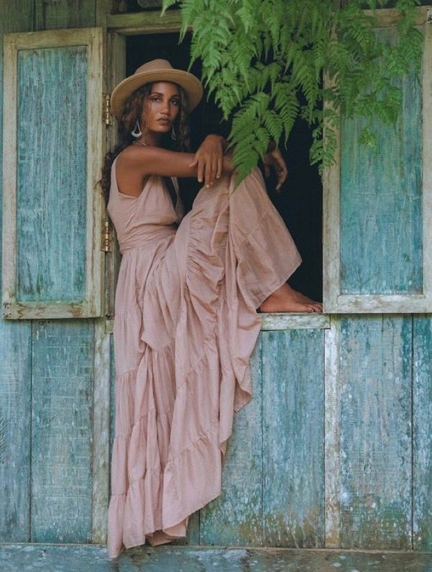 Woman in pink maxi dress and tan hat poses gracefully in a rustic teal window frame surrounded by ferns.