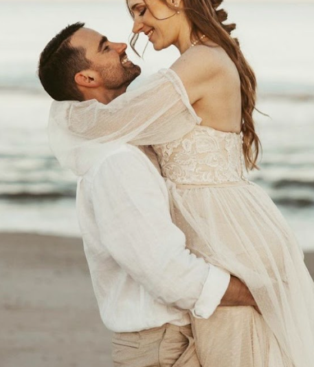 A romantic couple embraces on a beach during sunset, with the woman wearing a flowing white dress.