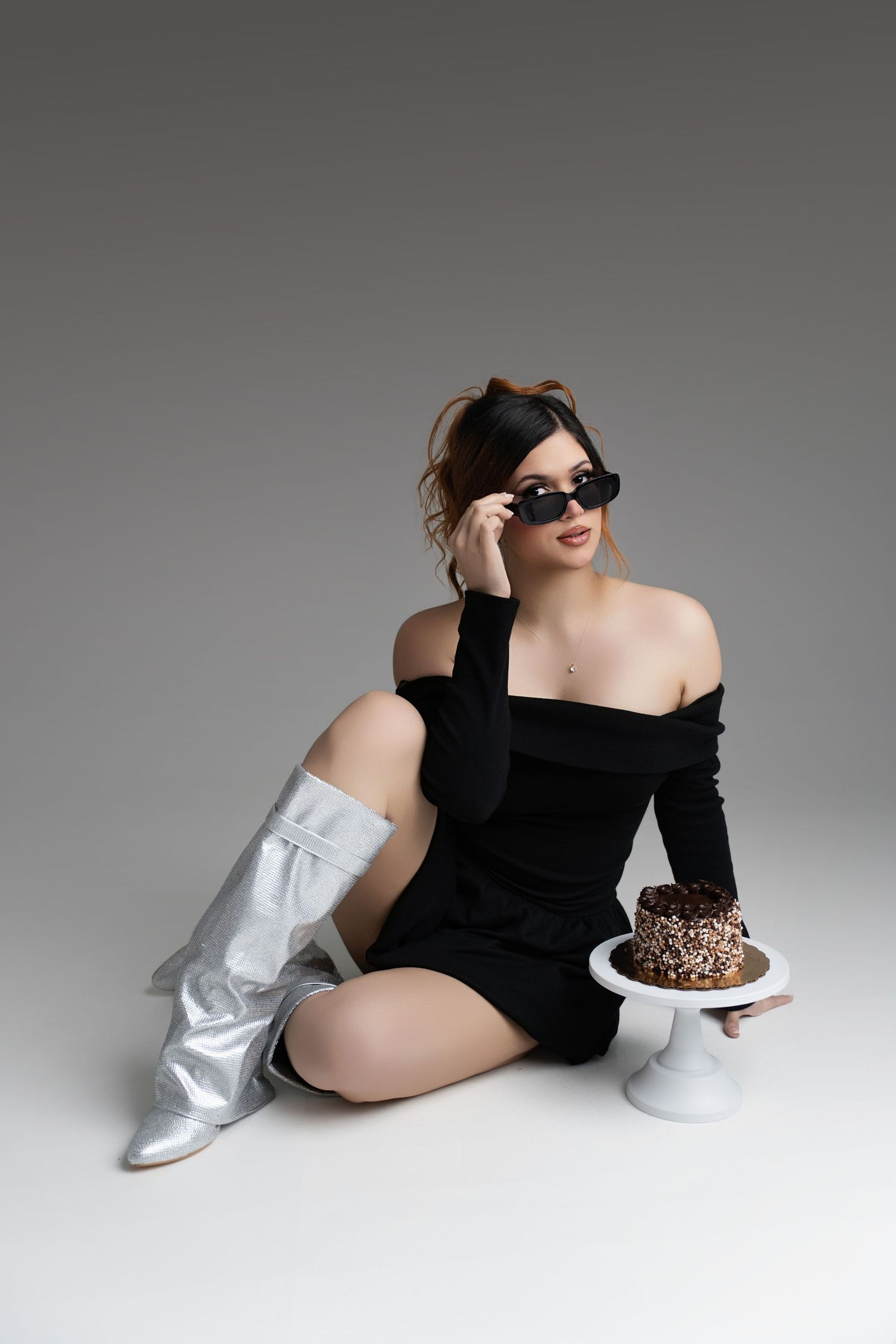 Fashion photo with model in black off-shoulder dress and silver boots posing with dessert on white pedestal against grey backdrop.
