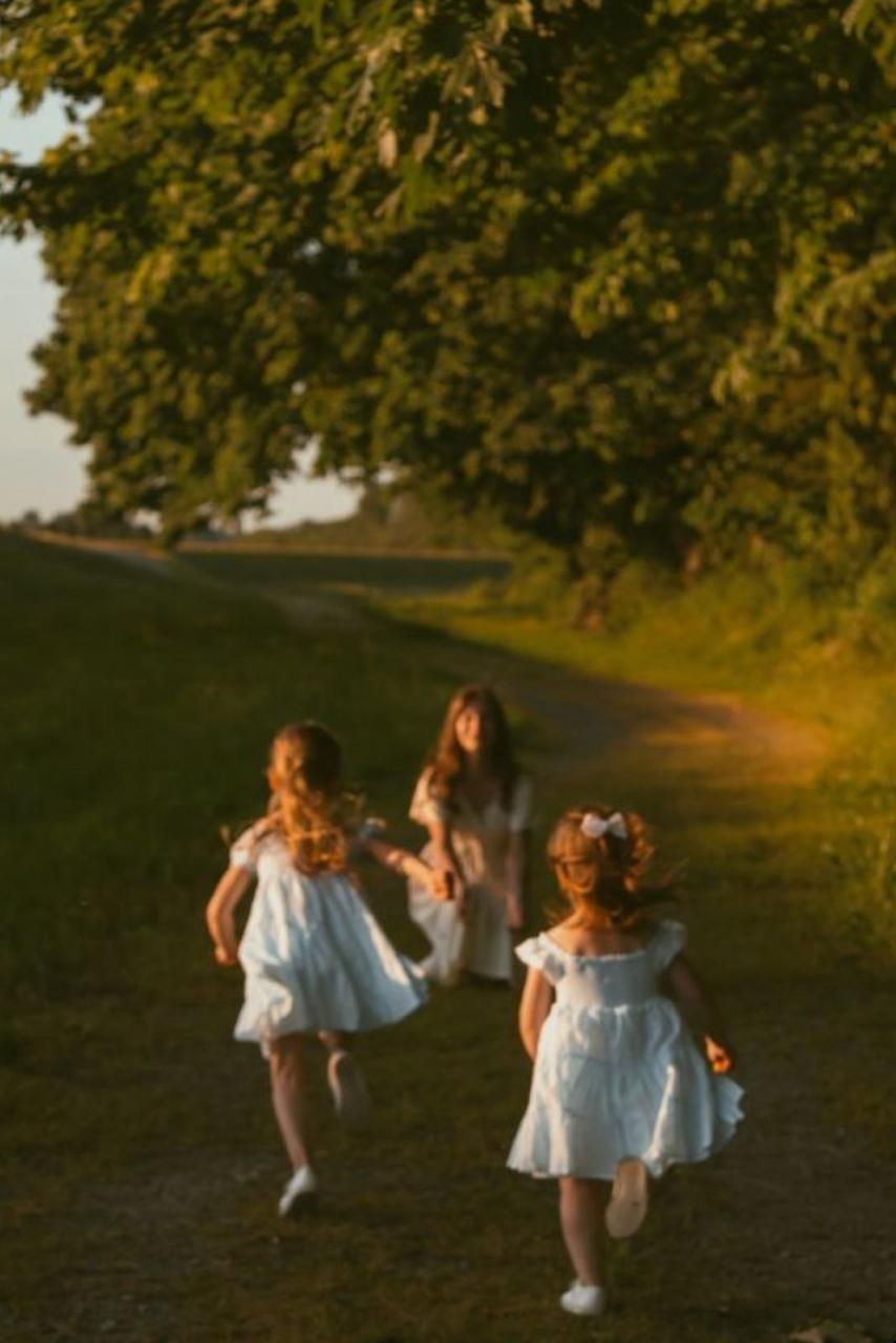 Children in white dresses running and playing together in a sunlit field with trees.