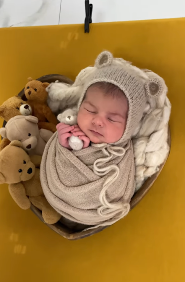 A sleeping baby swaddled in beige knit blanket lies in basket with small plush teddy bears on yellow background.