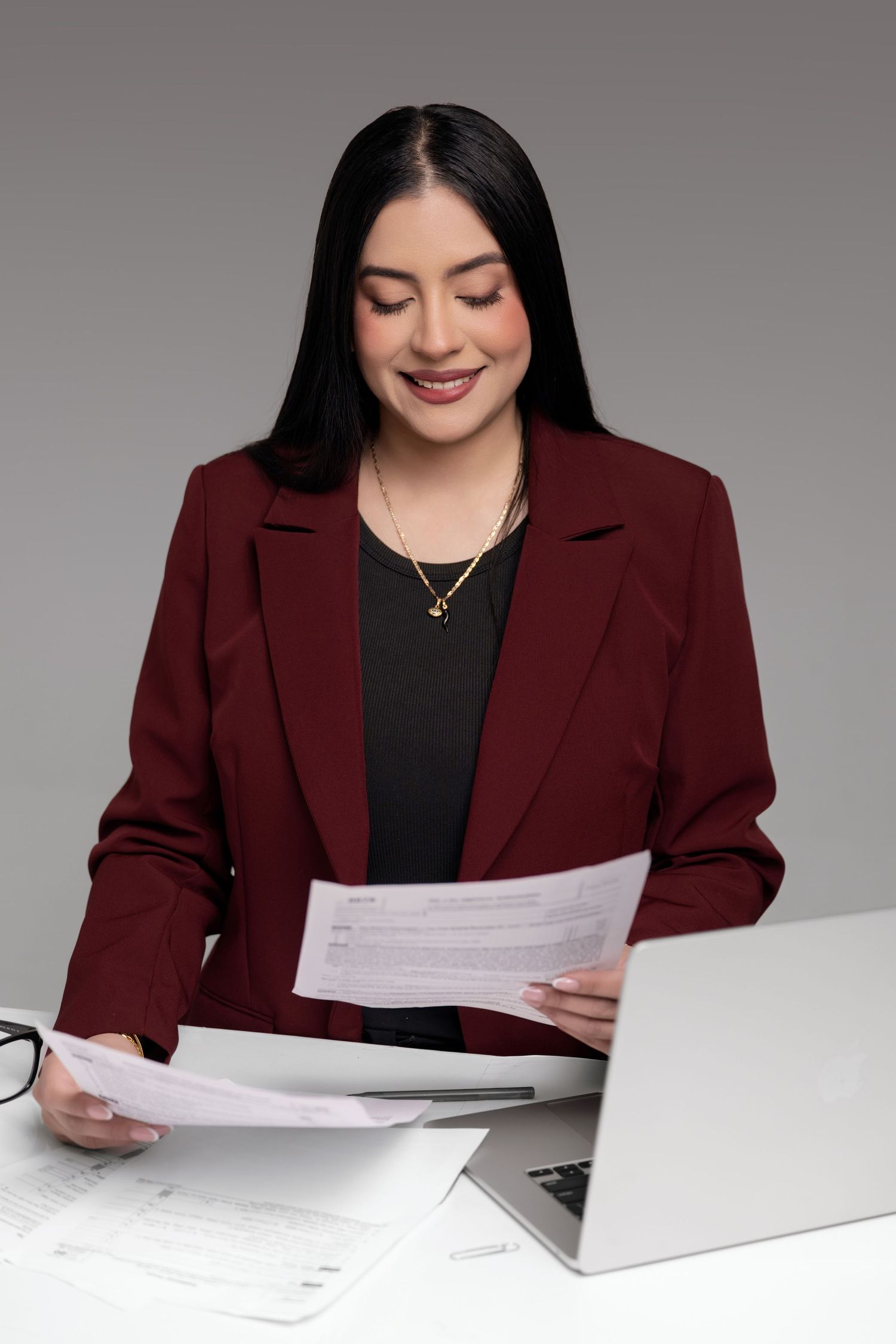 Professional in a burgundy blazer and black top reviews documents at a desk against a gray background.