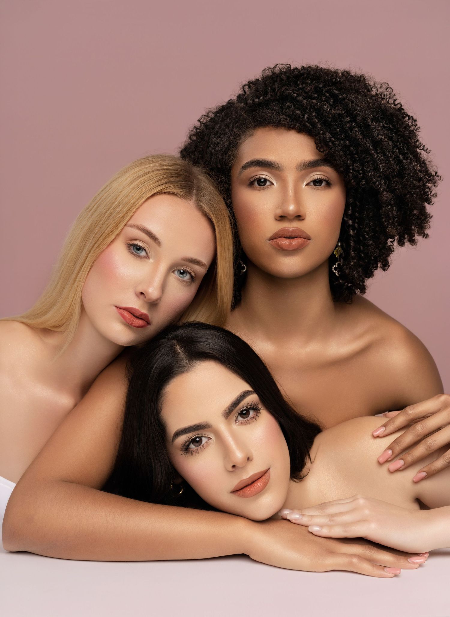 Three models with different hair colors and textures pose together in a beauty portrait against neutral background.