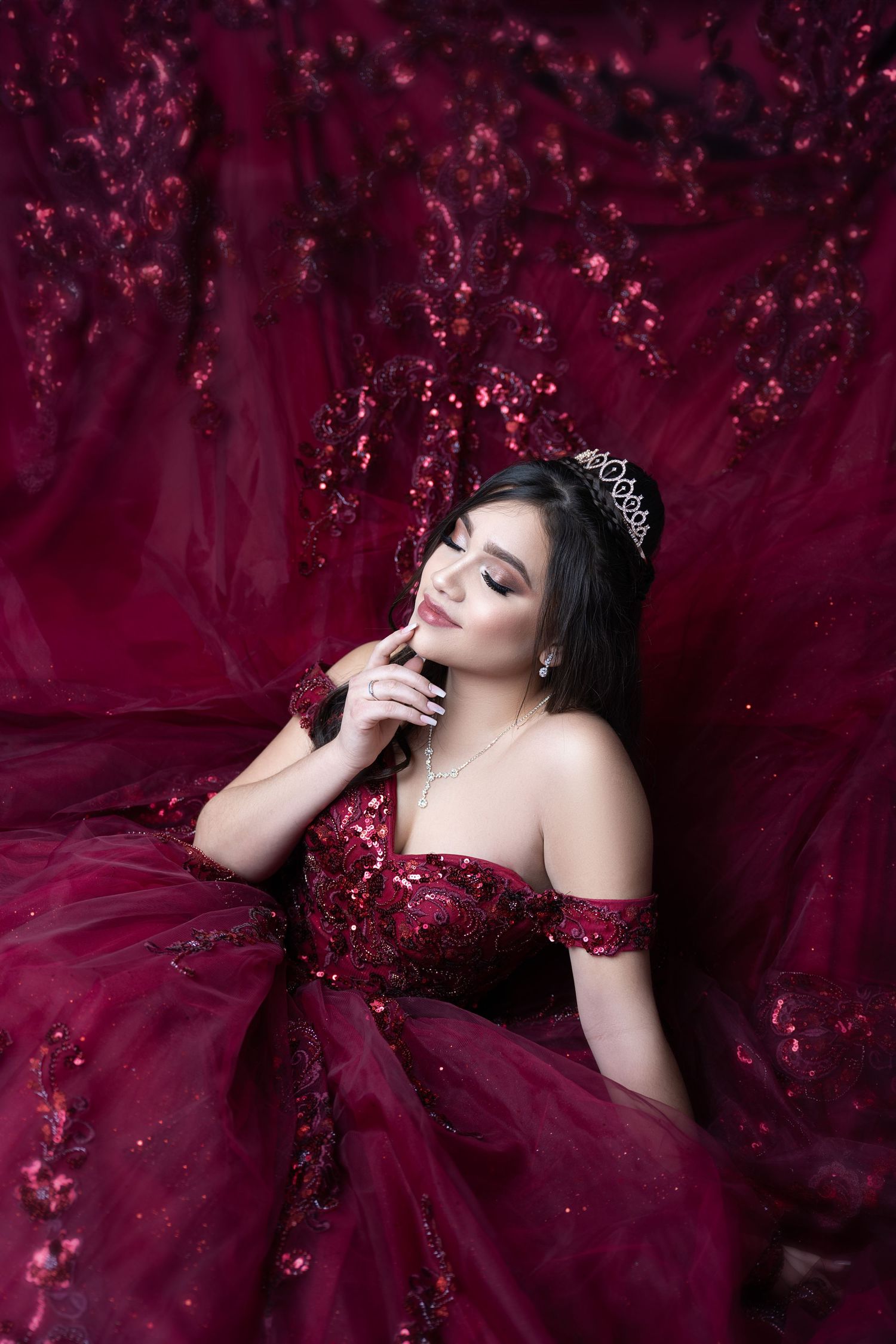 Young woman in a burgundy ball gown and tiara poses elegantly against a rich red floral backdrop.