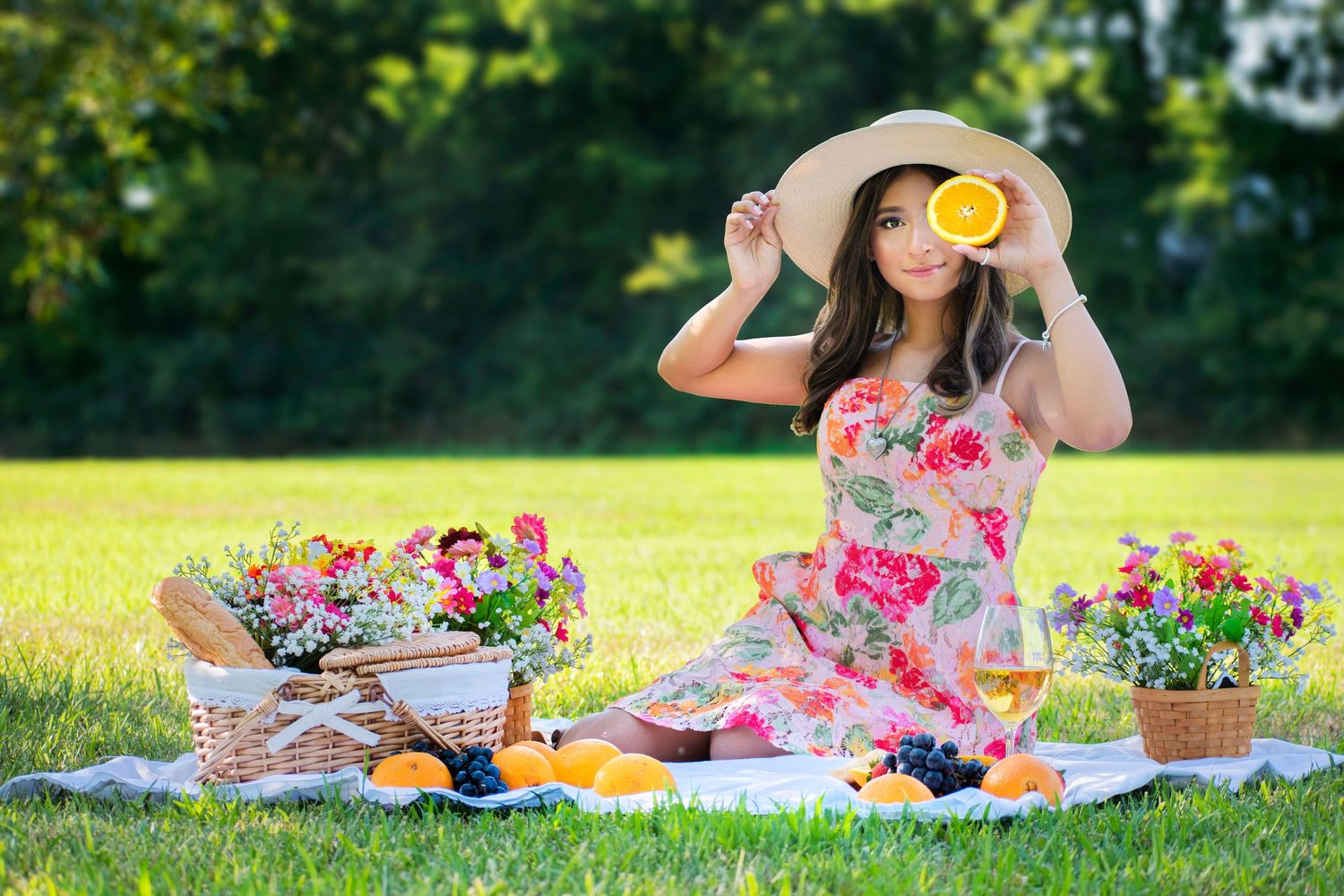 Young woman in floral dress and sun hat holds orange slice at a sunny outdoor picnic with flowers and fruit baskets on grass.