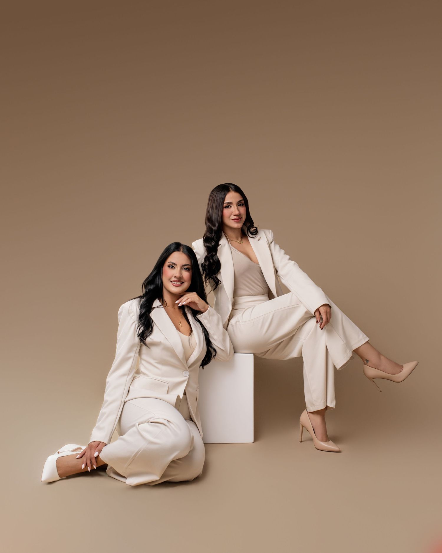 Two women in matching cream suits pose elegantly against a tan backdrop, one seated on floor, one on white cube.