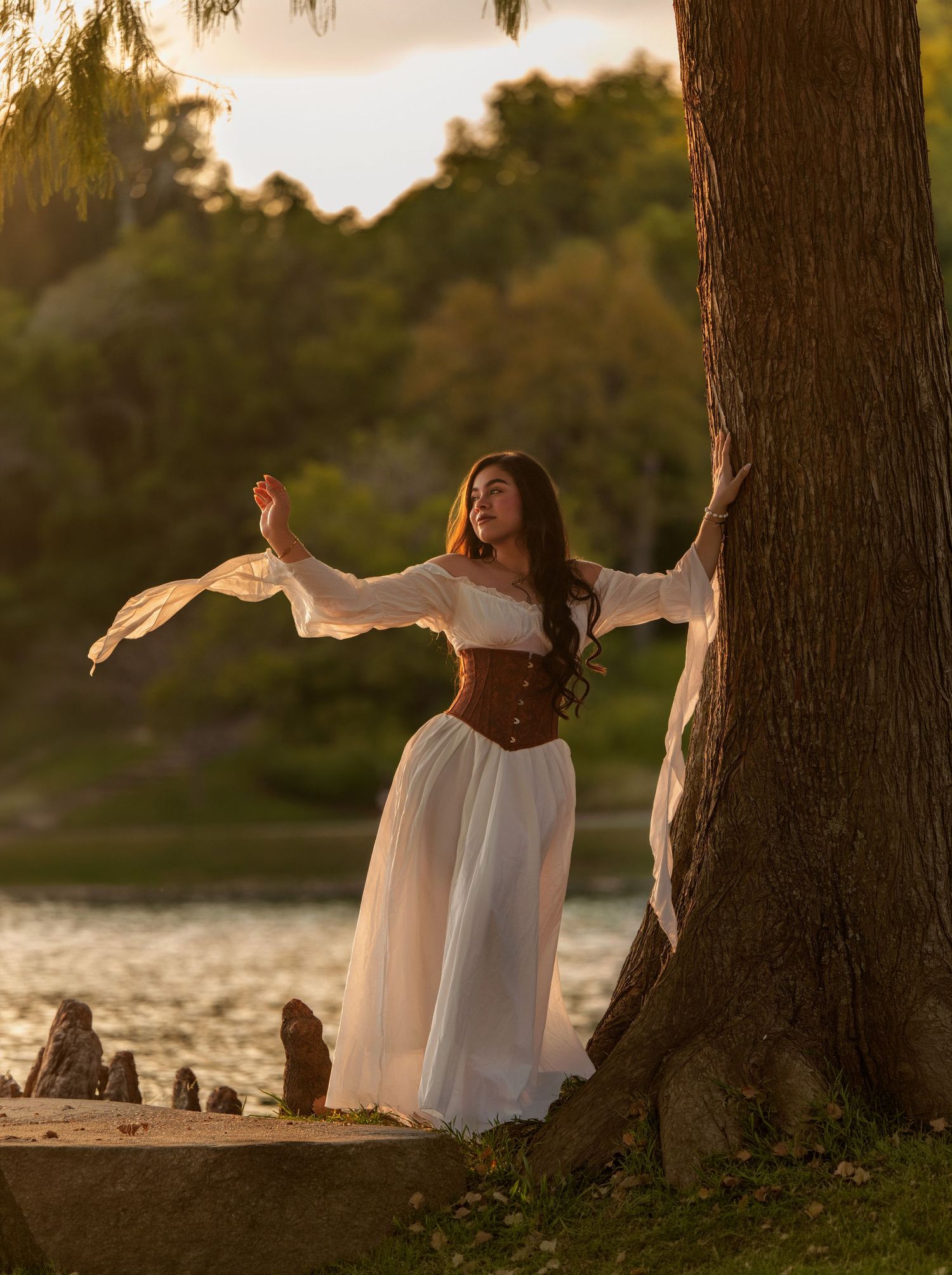 Woman in white dress and brown corset poses gracefully by a tree near a lake at golden hour.