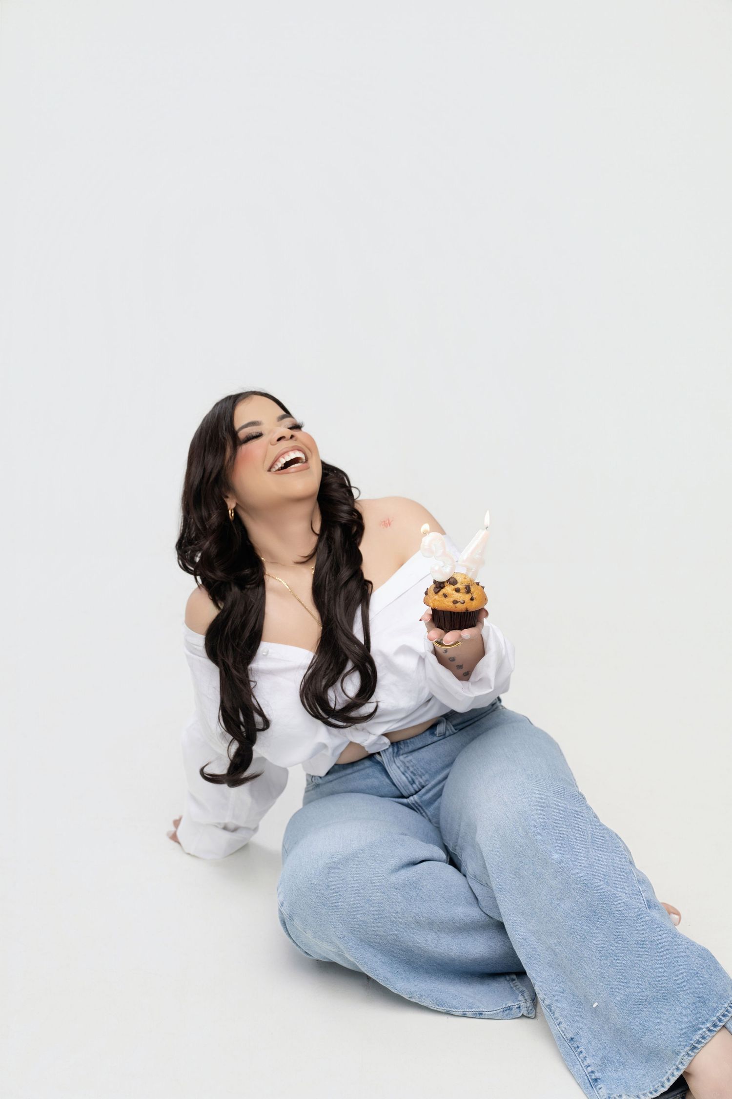 Smiling woman with long dark wavy hair laughs while sitting on a white studio floor holding a chocolate chip muffin with a lit candle.