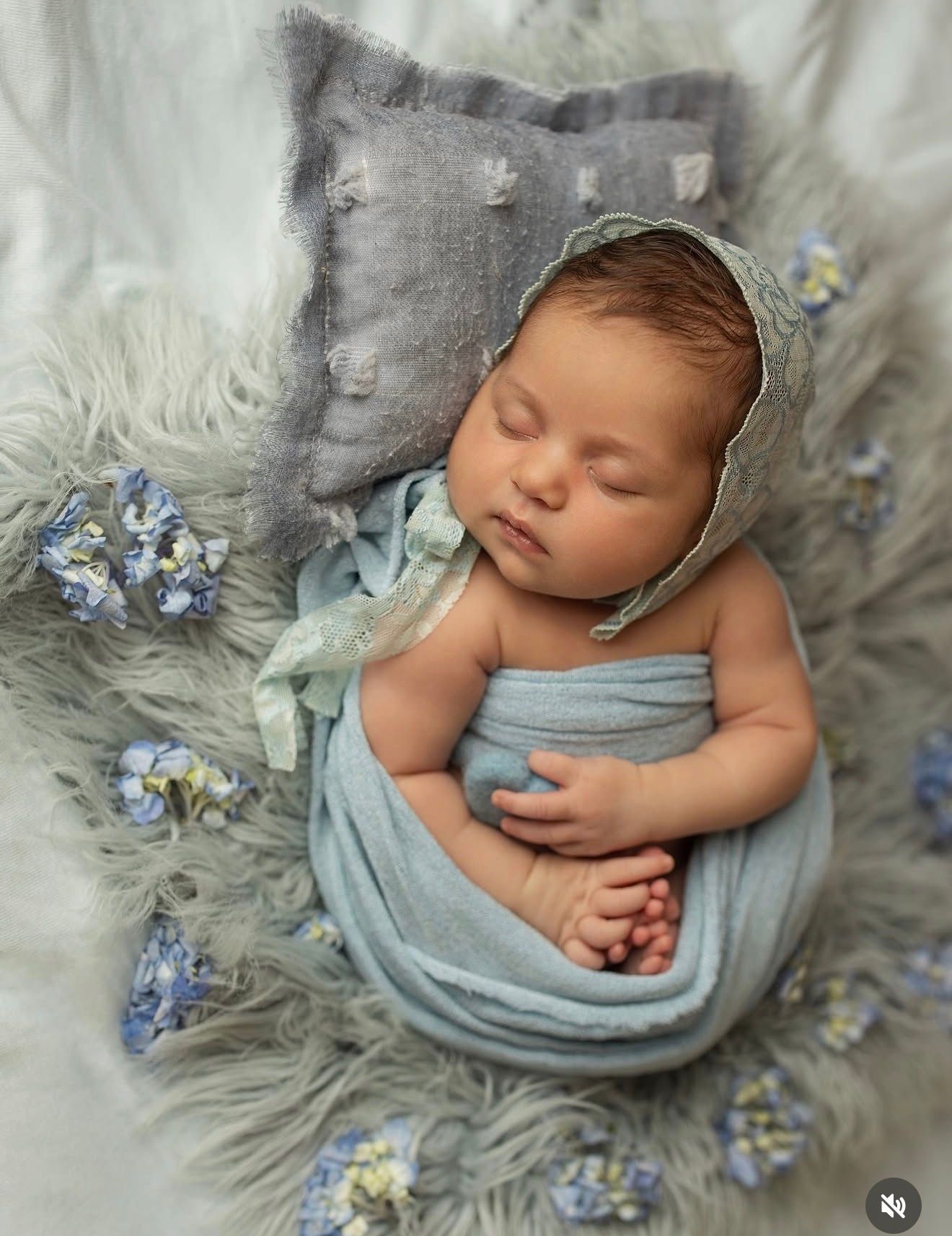 Newborn baby wrapped in light blue swaddle with bonnet, sleeping peacefully on fluffy gray fur with small blue flowers.