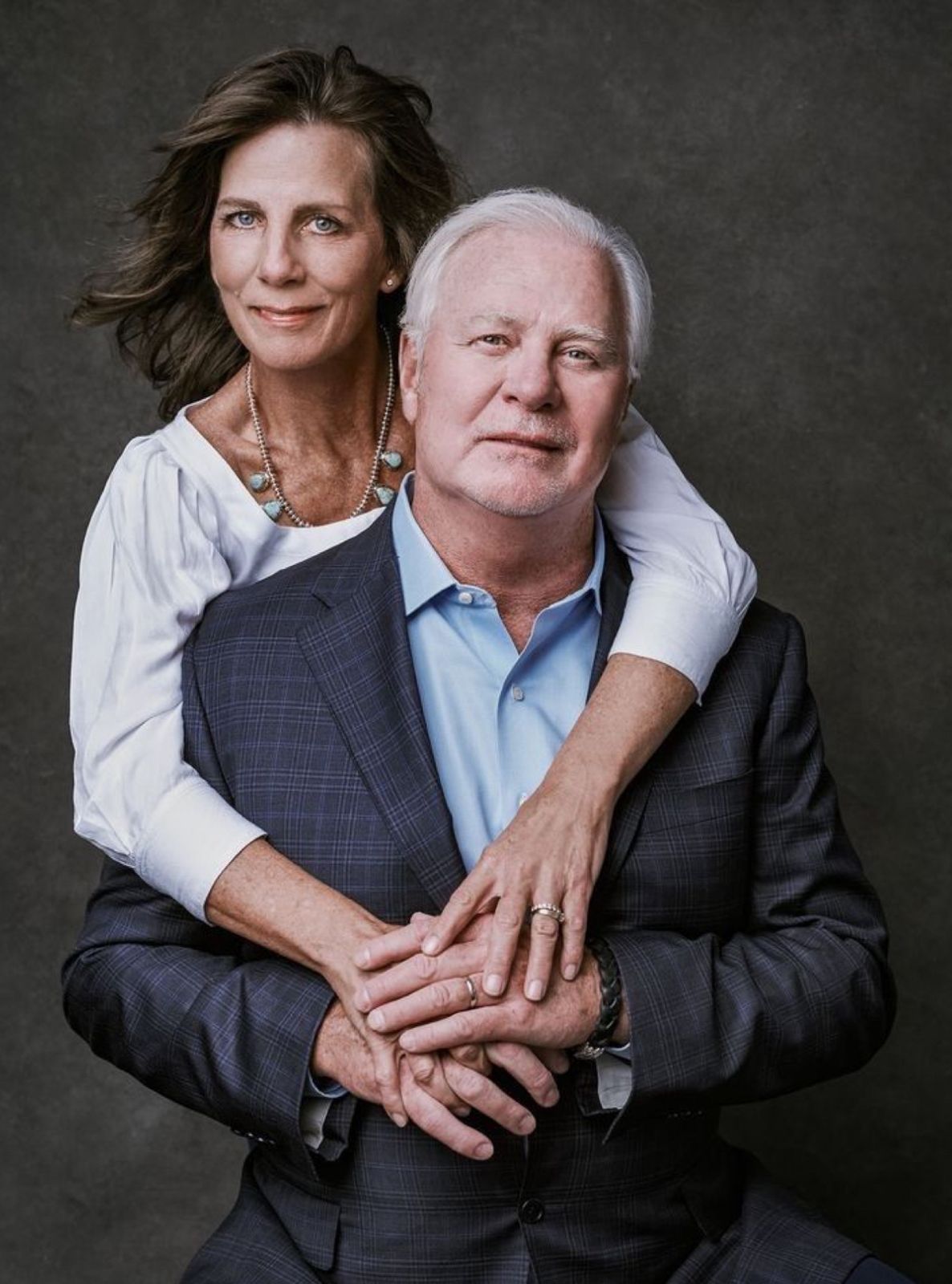 Mature couple posing affectionately, woman embracing man in blue suit from behind in professional studio portrait.
